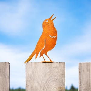 May include: An orange metal bird silhouette with an open beak, perched atop a wooden fence post. The bird has a simple, stylized design with a cut-out wing detail. The background is a bright blue sky with scattered clouds.