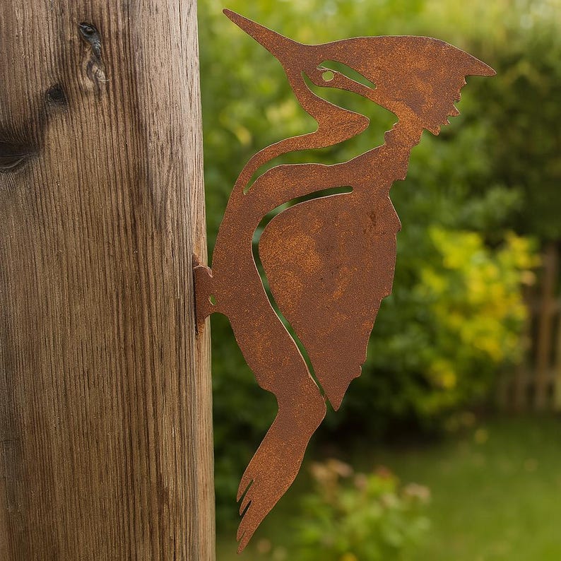 K&ouml;nnte beinhalten: Rostbraune Metall-Silhouette eines Spechts, befestigt an einem Holzpfosten. Das Profil des Vogels ist detailliert, mit einem langen Schnabel und strukturierten Federn. Der Hintergrund ist verschwommenes gr&uuml;nes Laub.