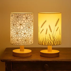 May include: Two table lamps with cylindrical shades. One shade has a floral pattern, the other depicts wheat stalks. Both lamps have wooden bases and stems, and are placed on a dark wooden surface. The lamps emit a warm, inviting glow.