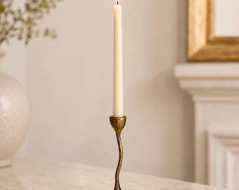 May include: A tall, slender, off-white taper candle is lit and held upright in a textured, antique-gold finished metal candlestick holder. The holder features a distinctively curved, organic stem that tapers down to a circular, flat base.