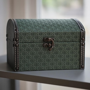 May include: A small, green decorative chest with a woven pattern. The chest has a curved top, dark brown leather trim, and a metal clasp. The edges are reinforced with small metal studs and white stitching.