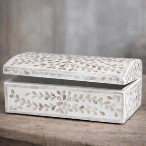 May include: A white decorative box with a curved lid, adorned with a mosaic leaf pattern in shades of beige, gray, and brown. The box is open, revealing a gray interior, and sits on a wooden surface.