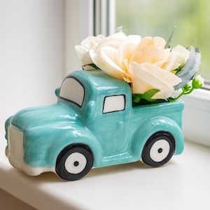 Puede incluir: Un jarrón de cerámica turquesa con forma de camión lleno de flores artificiales crema y blancas. El camión tiene ruedas negras con centros blancos y detalles blancos en la cabina. El jarrón está diseñado para arreglos florales.