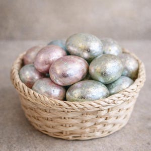 May include: A woven basket filled with decorative Easter eggs. The eggs have a metallic finish in shades of pink, silver, and blue. The basket is made of light brown woven material, and the eggs are oval-shaped.