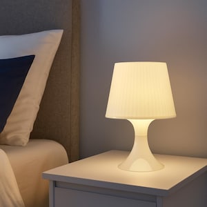 Könnte beinhalten: Eine weiße Nachttischlampe mit plissiertem Lampenschirm steht auf einem weißen Nachttisch. Die Lampe hat einen geschwungenen Fuß und strahlt ein warmes, weiches Licht aus. Ein Bett mit Kissen ist teilweise zu sehen.