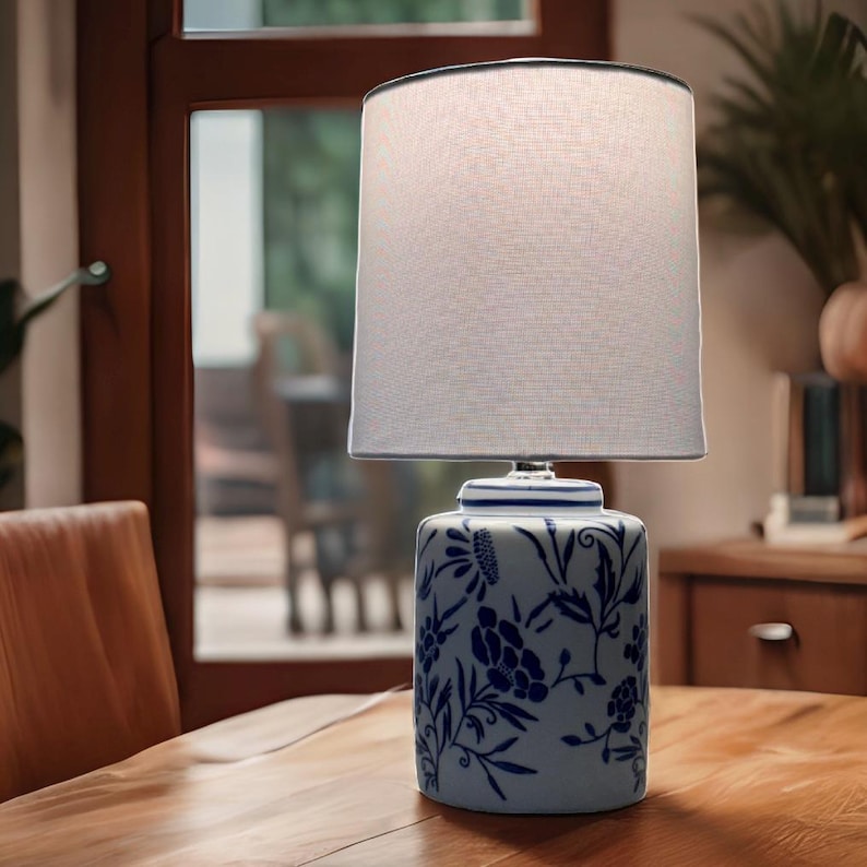 May include: A ceramic table lamp with a white lampshade and a blue and white floral design. The lamp sits on a wooden table. The lamp base has a cylindrical shape with a detailed floral pattern.