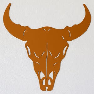 Könnte beinhalten: Ein rostfarbenes Metall-Wandkunstwerk in Form eines Bullenschädels. Der Schädel weist detaillierte Ausschnitte und geschwungene Hörner auf. Das Kunstwerk ist an einer weißen, strukturierten Wand befestigt und erzeugt einen auffälligen Kontrast.
