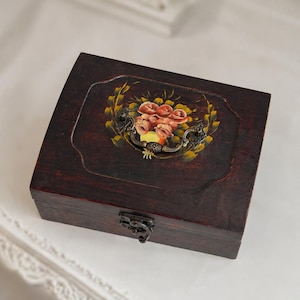 May include: A dark brown wooden box with a hinged lid and metal clasp. The lid features a painted floral design with pink roses, green leaves, and yellow accents. The box has a rectangular shape with a slightly curved top.