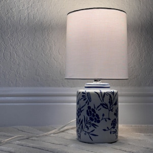 May include: A ceramic table lamp with a white lampshade and a blue and white floral design. The base of the lamp is cylindrical, and the lampshade is a simple, rounded shape. The lamp is plugged into an electrical outlet.