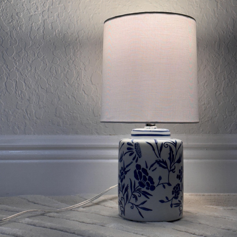 May include: A ceramic table lamp with a white lampshade and a blue and white floral design. The base of the lamp is cylindrical, and the lampshade is a simple, rounded shape. The lamp is plugged into an electrical outlet.