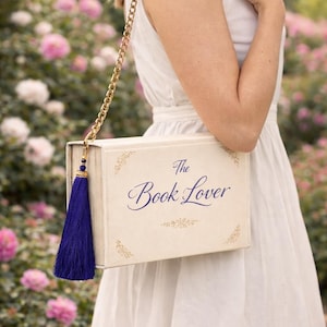 May include: Off-white book-shaped handbag with the text "The Book Lover" in navy blue script. The bag has a gold chain strap and a navy blue tassel. Floral accents are on the corners of the bag.