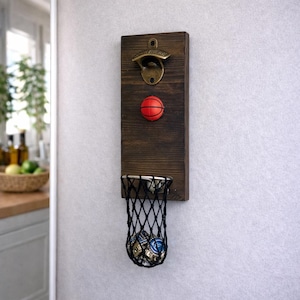 May include: A wooden bottle opener with a basketball theme. The dark brown wood has a bronze bottle opener at the top, a small red basketball, and a black net basket to catch bottle caps.