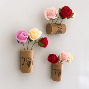 Può includere: Tre magneti decorativi, ciascuno con una base cilindrica in sughero stampata con una lettera J e un disegno a grappolo d'uva. Ogni sughero è sormontato da una piccola composizione di tre rose artificiali in schiuma nei toni rosa, rosso e crema.