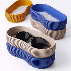 Peut inclure: Ensemble de trois plateaux ovales côtelés, jaune moutarde, bleu et beige. L'un des plateaux contient des lunettes de soleil noires. Les plateaux sont empilés, mettant en valeur leurs couleurs et textures contrastées. Intérieur gris clair.