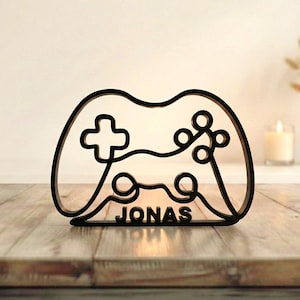 Peut inclure: Décoration noire en forme de manette de jeu en bois avec le nom "JONAS" en bas. La manette a une croix directionnelle, des boutons et un pavé directionnel. La décoration est sur une surface en bois.