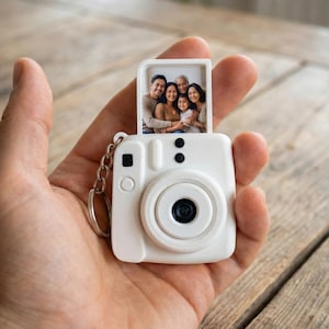 May include: A white camera-shaped keyring with a small photo of a family. The keyring has a black lens and a silver chain. The photo is of a family of five. The keyring is held in a hand.