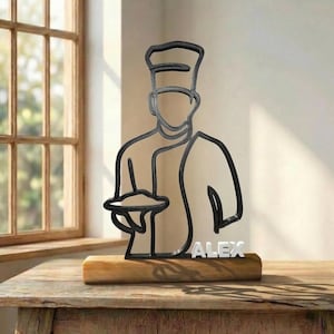 Puede incluir: Una escultura de arte lineal en metal negro de un chef con un gorro alto que sostiene una bandeja. La escultura está montada sobre una base de madera rectangular y presenta el nombre ALEX en letras blancas de bloque en la base de la figura.