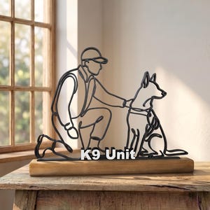 Peut inclure: Une sculpture en fil de fer noir représentant une personne agenouillée à côté d'un chien, montée sur une base en bois rectangulaire. La sculpture comporte le texte K9 Unit en lettres blanches à la base entre les figures.