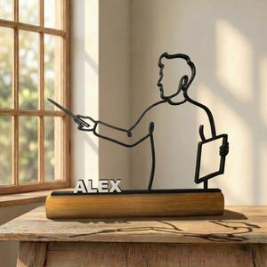 Peut inclure: Une sculpture en fil de métal noir représentant une silhouette d'enseignant tenant un pointeur et un livre, fixée sur une base cylindrique en bois. Le nom ALEX est affiché en lettres capitales argentées sur la base, devant la silhouette.