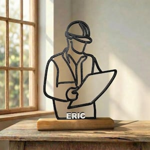 Peut inclure: Une sculpture en fil de fer noir représentant un ouvrier du bâtiment portant un casque et un gilet, tenant un plan. La silhouette est fixée sur une base rectangulaire en bois avec le nom ERIC en lettres capitales blanches en bas.