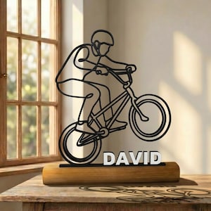 Puede incluir: Una escultura de alambre negro que representa a una persona montando una bicicleta en posición de salto, montada sobre una base rectangular de madera. El nombre DAVID aparece en letras blancas de bloque en la base de la escultura.