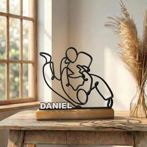 Op de afbeelding: Een zwart draadsculptuur dat twee figuren in een worstelgreep uitbeeldt, gemonteerd op een rechthoekige houten voet. De naam DANIEL staat in witte blokletters op de voorzijde van de voet, onder het metalen draadkunstwerk geplaatst.