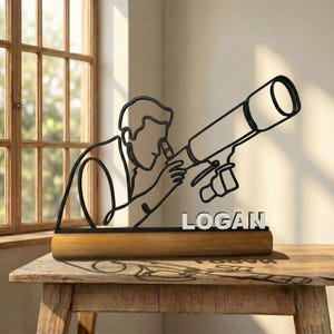 Peut inclure: Une sculpture en fil de fer noir représentant une personne regardant à travers un télescope, montée sur une base cylindrique en bois. Le nom LOGAN est affiché en lettres capitales blanches sur le devant de la base, sous la silhouette.