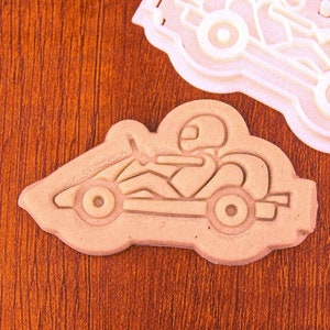 Peut inclure: Deux emporte-pièces en forme de karting et de pilote. L'un est de couleur beige clair, l'autre blanc. Le design du karting comprend le pilote, le casque et les roues. Les emporte-pièces sont sur une surface en bois brun.