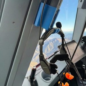 Peut inclure: Casque d'aviation avec un support bleu pour un appareil et des bouchons d'oreille orange. Le casque est noir et gris avec la marque "BOSE" visible. L'arrière-plan montre une vue depuis un cockpit d'avion.