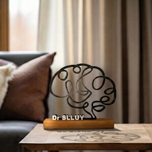 Puede incluir: Una escultura de alambre de metal negro con forma de cerebro humano, montada sobre una base rectangular de madera. La base presenta el texto blanco Dr SLLUY. La escultura est&aacute; compuesta por una l&iacute;nea continua que forma el cerebro.