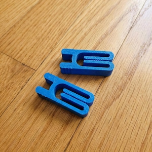 Peut inclure: Deux clips en plastique bleu vif, imprimés en 3D. Chaque clip a une forme unique avec une section centrale et deux broches. Les clips reposent sur une surface en bois avec un motif de grain de bois naturel.