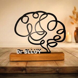 Puede incluir: Una escultura de l&iacute;nea continua negra con forma de cerebro humano, montada sobre una base rectangular de madera. El texto blanco Dr SLLUY est&aacute; fijado en la parte frontal de la base, posicionado directamente bajo la estructura cerebral.