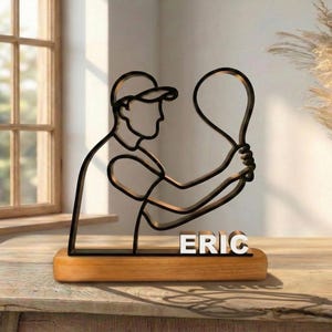 May include: A black metal wire-frame sculpture of a tennis player wearing a cap and holding a racket, mounted on a rectangular wooden base. The name ERIC is displayed in white block letters on the front of the base below the tennis player.