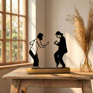 May include: A black metal sculpture of two figures in motion, mounted on a wooden base. One figure points, the other appears to be dancing. The sculpture is placed on a wooden table near a window.