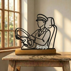 Könnte beinhalten: Eine schwarze Metalldrahtskulptur, die einen Fahrer mit Mütze auf einem Sitz zeigt, der ein Lenkrad hält. Der Name Sepp ist in weißen Blockbuchstaben an der Basis der Figur angebracht, die auf einem rechteckigen Holzblock montiert ist.