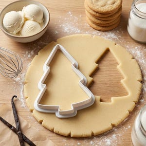 Peut inclure: Un emporte-pièce en plastique blanc en forme de sapin de Noël sur une pâte à biscuits étalée. L'image montre également un bol de glace à la vanille, une pile de biscuits et un pot de sucre.