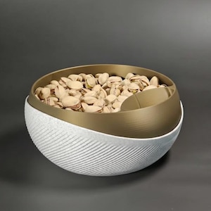 Peut inclure: Un bol décoratif rempli de pistaches. Le bol a une base blanche texturée avec un intérieur et un bord de couleur or. Les pistaches sont brun clair et remplissent le bol.