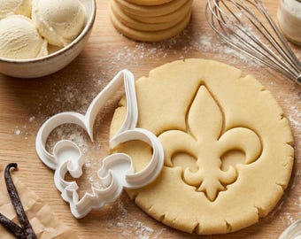 Könnte beinhalten: Ein weißer Ausstecher in Form einer Fleur-de-Lis neben einem ausgestochenen Keks auf einer Holzoberfläche. Vanilleschoten, eine Schüssel Vanilleeis und ein Stapel Kekse sind ebenfalls zu sehen. Ein Schneebesen und Zucker befinden sich im Hintergrund.