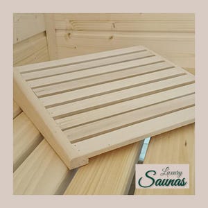 Può includere: Una panca per sauna in legno chiaro con piano a doghe. La panca è rettangolare con bordi arrotondati ed è realizzata in legno naturale. L'immagine mostra la panca in un ambiente sauna, con il testo "Luxury Saunas" in basso a destra.