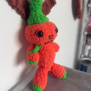 Puede incluir: Una muñeca de ganchillo naranja hecha a mano con un sombrero, bufanda y detalles verdes. La muñeca tiene ojos negros y una boca cosida. La muñeca está sobre una superficie gris.