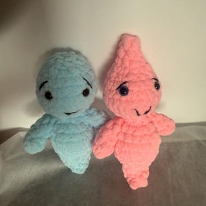 Puede incluir: Dos muñecos de ganchillo hechos a mano. Uno es azul claro, el otro rosa. Ambos muñecos tienen ojos negros y pequeñas sonrisas. Los muñecos tienen una forma de cuerpo redondeada con brazos y piernas pequeños. Los muñecos están hechos de hilo suave y texturizado.