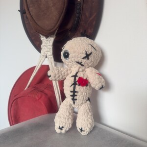 Puede incluir: Una muñeca vudú hecha a mano, de color crema, con detalles cosidos en negro y un corazón rojo. La muñeca tiene ojos negros en forma de cruz y está de pie frente a un sombrero de vaquero marrón y una bolsa roja.