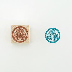 Puede incluir: Un sello de madera con un diseño de tinta marrón de tres hojas estilizadas dentro de un círculo. Una impresión en tinta turquesa del mismo diseño está al lado del sello. El sello está sobre un fondo blanco.