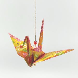 May include: Origami crane ornament in shades of orange, yellow, and pink with floral patterns. The paper crane is suspended by a gold chain with a red bead accent. A traditional Japanese craft.