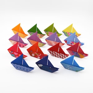 Puede incluir: Una colección de coloridos barcos de origami, cada uno doblado con papel estampado. Los barcos presentan tonos vibrantes de naranja, verde, morado, rojo y azul, con diseños intrincados en las velas y los cascos. Los barcos de papel están dispuestos sobre una superficie blanca.