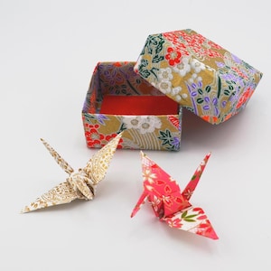 May include: An open, floral-patterned box with a red interior and a matching lid. Two origami cranes, one gold and one pink, are positioned near the box. The box and cranes are on a white surface. The box and cranes are decorative.