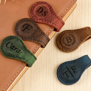 May include: A collection of personalized leather bookmarks in various colors, including red, brown, green, and blue. Each bookmark features a unique initial or name engraving. The bookmarks are arranged on a leather notebook.
