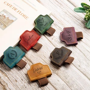 May include: Six leather pen holders with elastic bands in green, red, teal, yellow, brown, and purple. Each holder features unique laser-engraved designs including names like Sophia and Gabriel, initials like A, F, and B&F, and a dove icon.