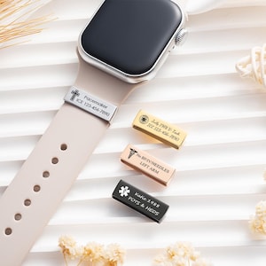 May include: A beige smartwatch band featuring a silver medical alert tag engraved with a cross and the text Pacemaker ICE 123-456-7890. Three additional rectangular medical alert tags in gold, rose gold, and black are displayed nearby.