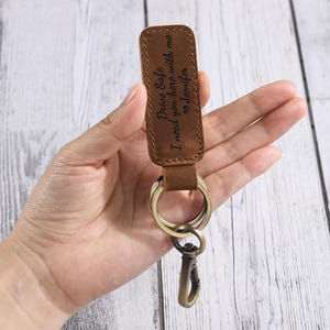 May include: A brown leather keychain with a metal clasp and key rings. The keychain has the engraved text "Drive Safe I need you here with me Jennifer". The keychain is held in a hand.